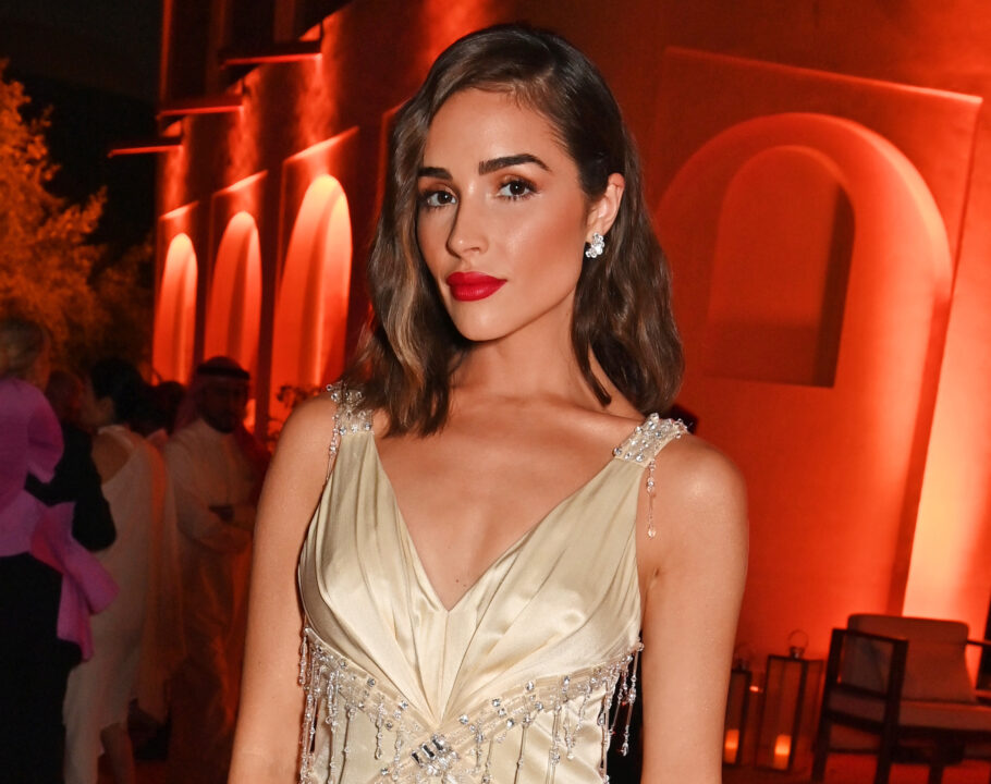 Olivia Culpo wears a gold dress and red lipstick in a red-lit ballroom